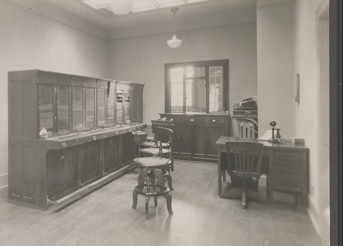 A telephone exchange room around 1900, with a long wooden switchboard, operator stools and a desk.